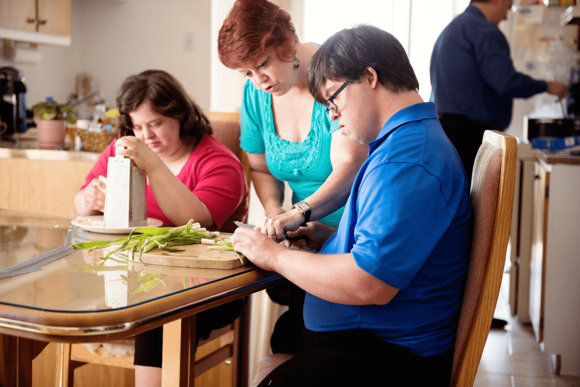 Woman guiding two young adults while they prepare vegetables at the kitchen table. Alt
